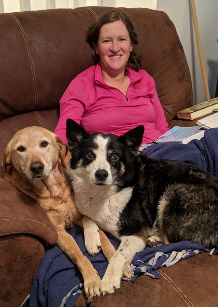 A woman is sitting on a recliner with two dogs and a book.
